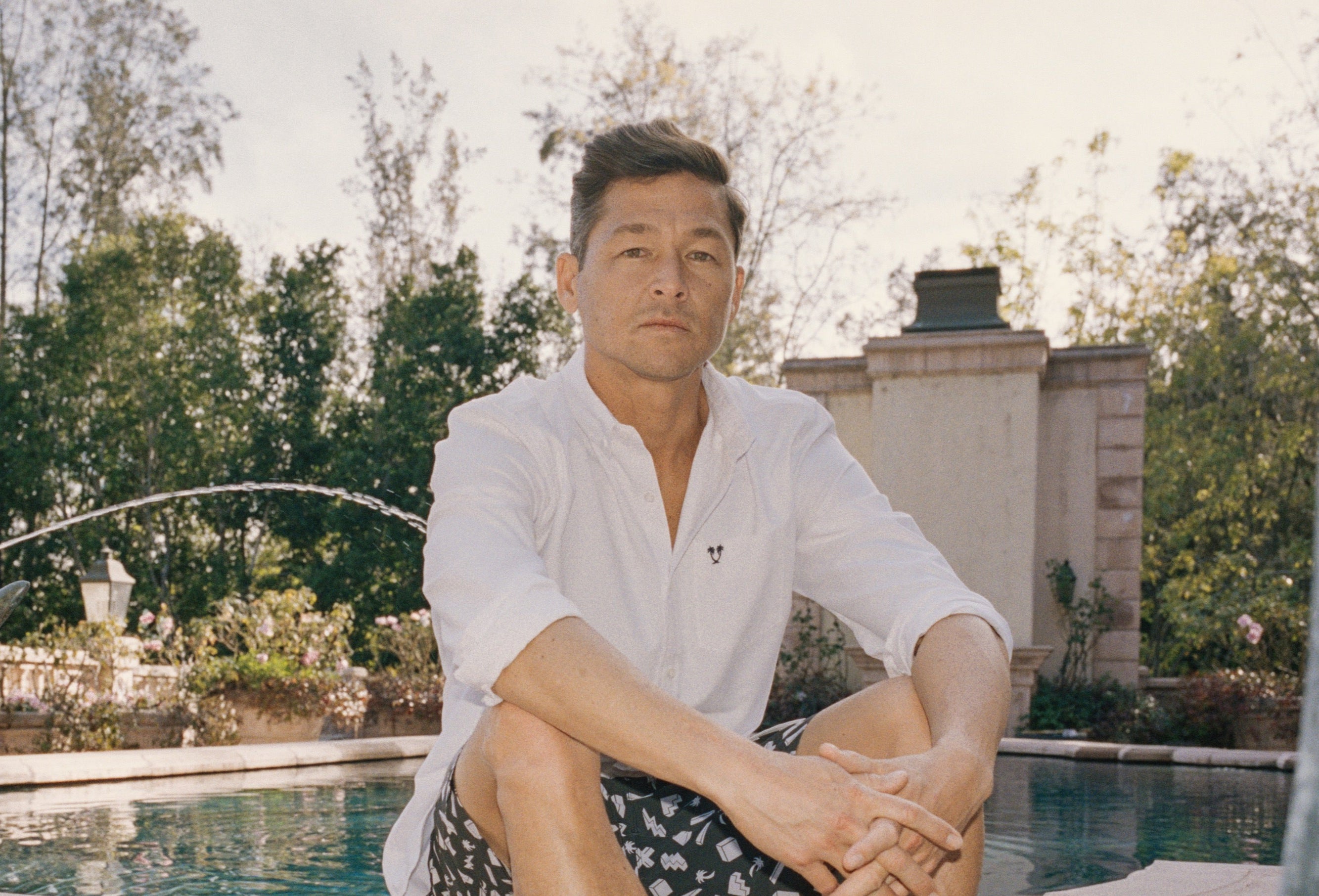 Man sitting by a pool with a garden and fountain in the background
